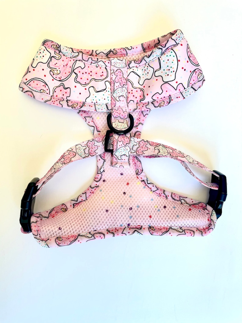 May include: Pink dog harness with a cookie pattern. The harness has a mesh lining and adjustable straps with black buckles.