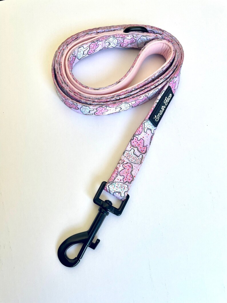 May include: Pink dog leash with a white background and a pattern of pink and white cookies. The leash has a black clip and the text "Smash Face" is printed on the leash.