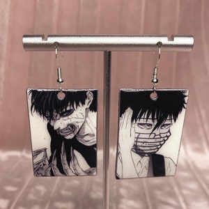 Puede incluir: Un par de pendientes rectangulares en blanco y negro con un personaje de anime. Los pendientes presentan una imagen en blanco y negro de un personaje con cabello negro y una máscara blanca. Los pendientes cuelgan de ganchos plateados.