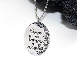 May include: Silver necklace with an oval pendant that says "live love aloha" with a small heart symbol between "live" and "love". The pendant is surrounded by a border of small flower designs.