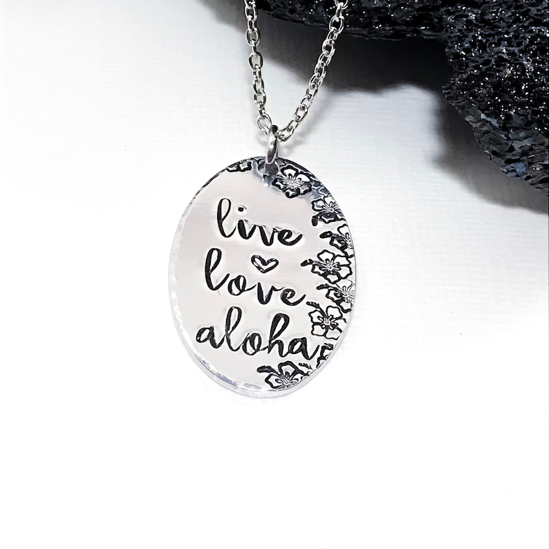 May include: Silver necklace with an oval pendant that says "live love aloha" with a small heart symbol between "live" and "love". The pendant is surrounded by a border of small flower designs.