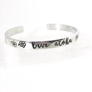 May include: Silver bangle bracelet with the words "Live Aloha" engraved on it. The bracelet is decorated with two small flower designs.