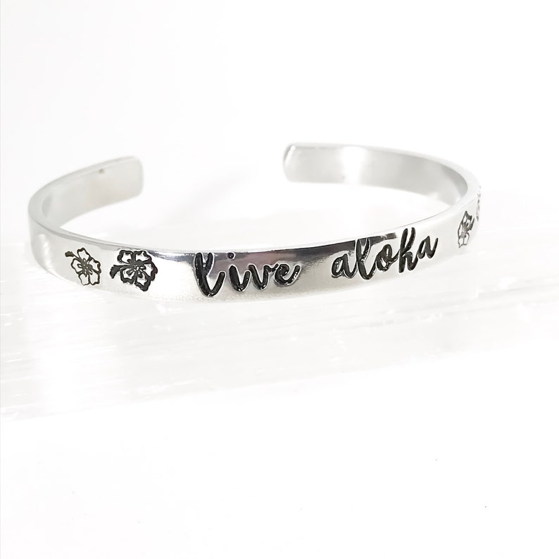 May include: Silver bangle bracelet with the words "Live Aloha" engraved on it. The bracelet is decorated with two small flower designs.