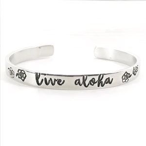May include: Silver bangle bracelet with the words "live aloha" engraved on it. The bracelet is decorated with small flower designs.