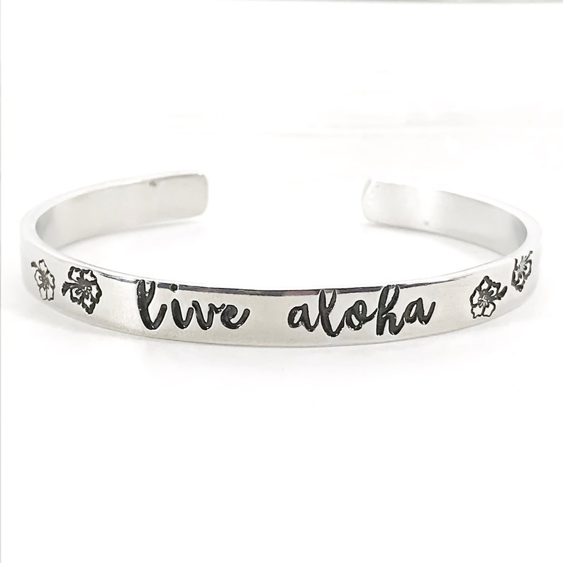 May include: Silver bangle bracelet with the words "live aloha" engraved on it. The bracelet is decorated with small flower designs.