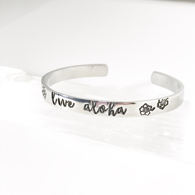 May include: Silver bracelet with the words "live aloha" engraved on it. Two small flower designs are also engraved on the bracelet.