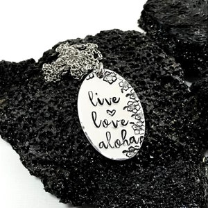 May include: Silver necklace with an oval pendant that says "live love aloha" with a small heart between "live" and "love". The pendant is surrounded by a border of small flowers.