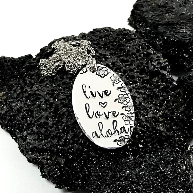 May include: Silver necklace with an oval pendant that says "live love aloha" with a small heart between "live" and "love". The pendant is surrounded by a border of small flowers.