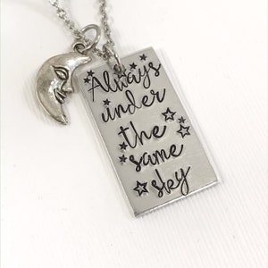 May include: Silver necklace with a crescent moon charm and a rectangular tag that reads "Always under the same sky" with stars around the text.