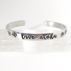 May include: Silver bangle bracelet with the words "live aloha" engraved on it. The bracelet is decorated with two small flower designs.