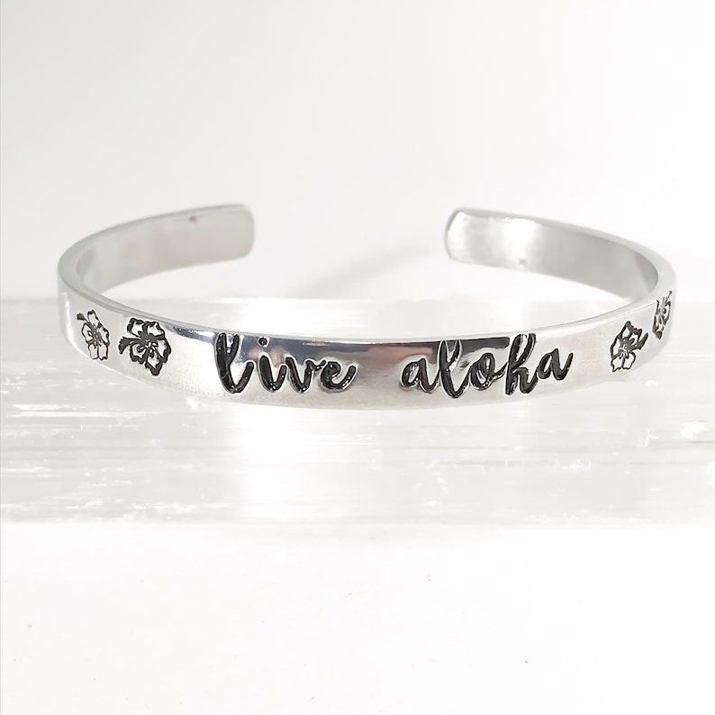 May include: Silver bangle bracelet with the words "live aloha" engraved on it. The bracelet is decorated with two small flower designs.