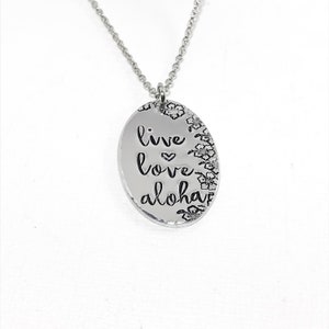 May include: Silver necklace with an oval pendant that says "live love aloha" with a small heart between "live" and "love". The pendant is surrounded by small flower designs.