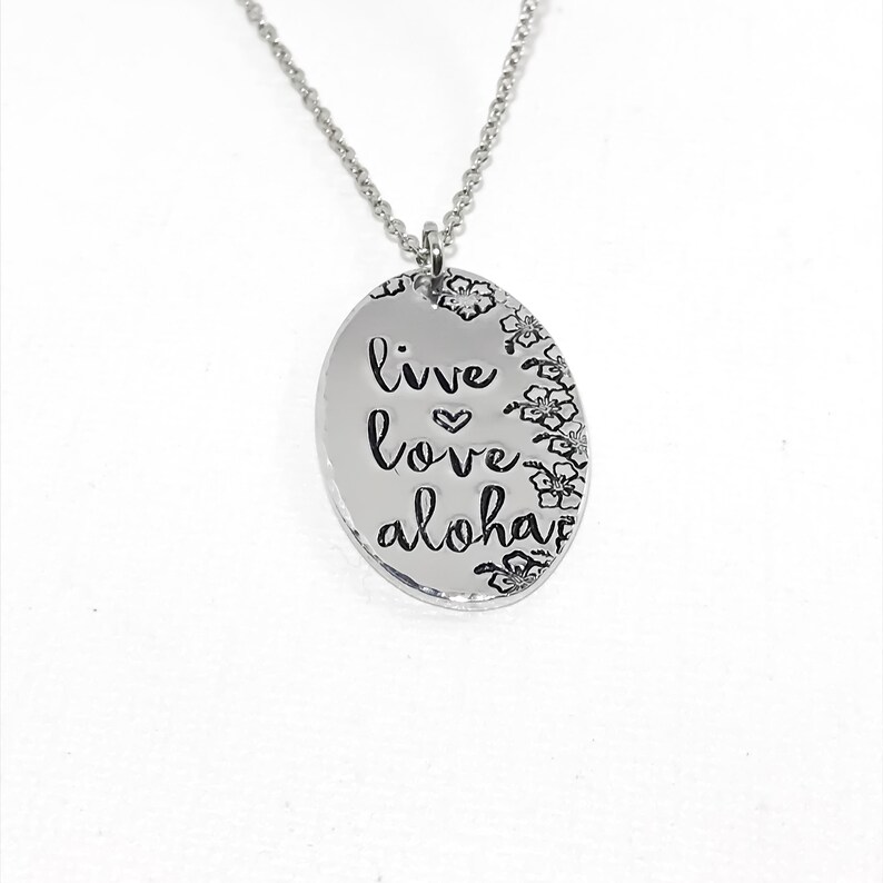 May include: Silver necklace with an oval pendant that says "live love aloha" with a small heart between "live" and "love". The pendant is surrounded by small flower designs.