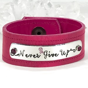 May include: Pink leather bracelet with a silver plate engraved with the words "Never Give Up" and a pink ribbon symbol. The bracelet has two small pink gemstones.