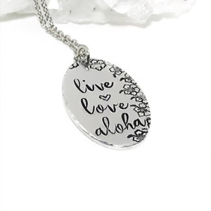 May include: Silver necklace with an oval pendant that says "live love aloha" with a small heart symbol between "love" and "aloha". The pendant is surrounded by a border of small flower designs.