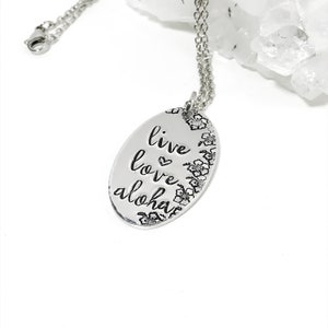 May include: Silver necklace with an oval pendant that says "live love aloha" with a heart symbol between the words "live" and "love". The pendant is surrounded by small flower designs.