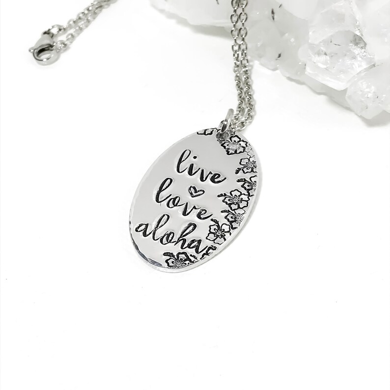May include: Silver necklace with an oval pendant that says "live love aloha" with a heart symbol between the words "live" and "love". The pendant is surrounded by small flower designs.