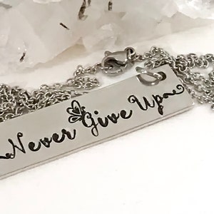 May include: A silver necklace with a rectangular pendant that says "Never Give Up" in a cursive font. The pendant has a small butterfly design on the left side.
