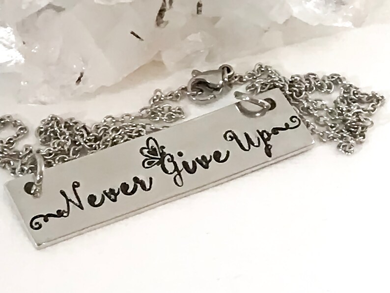 May include: A silver necklace with a rectangular pendant that says "Never Give Up" in a cursive font. The pendant has a small butterfly design on the left side.
