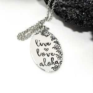 May include: Silver necklace with an oval pendant that says "live love aloha" with a small heart between "love" and "aloha". The pendant is surrounded by a floral design.