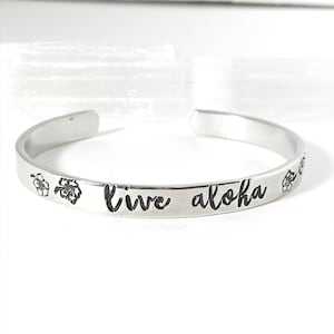May include: Silver bangle bracelet with the words "live aloha" engraved on it. Two small flower designs are on either side of the text.