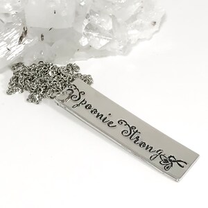 May include: A silver necklace with a rectangular pendant that reads "Spoonie Strong" in a cursive font.