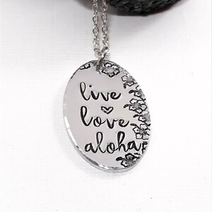 May include: Silver necklace with an oval pendant that says "live love aloha" with a small heart between "live" and "love". The pendant is surrounded by a border of small flower designs.