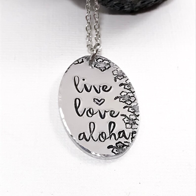 May include: Silver necklace with an oval pendant that says "live love aloha" with a small heart between "live" and "love". The pendant is surrounded by a border of small flower designs.