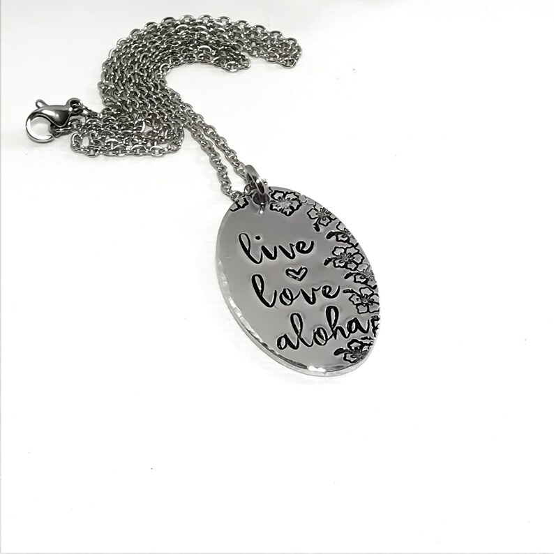 May include: Silver necklace with an oval pendant that says "live love aloha" with a small heart between "live" and "love". The pendant is surrounded by a floral design.