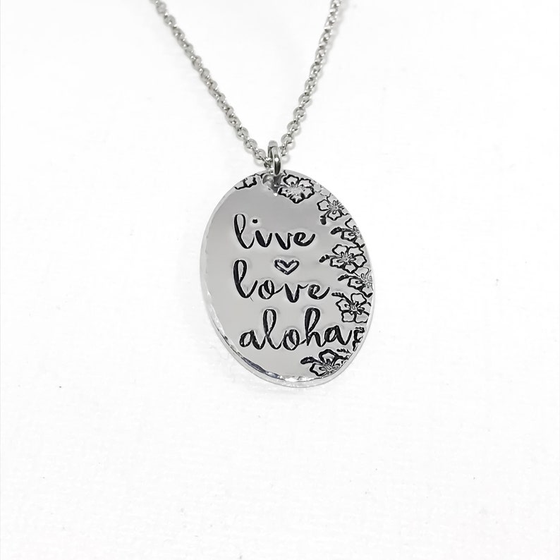 May include: Silver necklace with an oval pendant that says "live love aloha" with a small heart between the words "live" and "love". The pendant is surrounded by small flower designs.