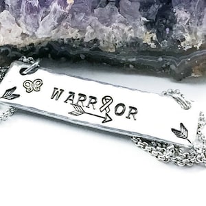 May include: Silver necklace with a rectangular pendant that says "WARRIOR" with an arrow and a ribbon symbol. The pendant also has a small butterfly charm.