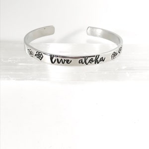 May include: A silver bangle bracelet with the words "Live Aloha" engraved on it. The bracelet is decorated with small flower designs.