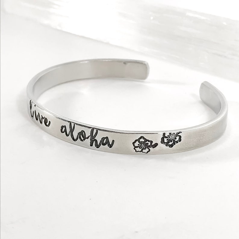 May include: Silver bangle bracelet with the words "live aloha" engraved on it. Two small hibiscus flowers are also engraved on the bracelet.