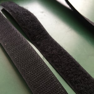 Könnte beinhalten: Nahaufnahme von schwarzen Klettverschlussstreifen. Die Streifen bestehen aus gewebtem Nylon und haben eine strukturierte Oberfläche. Ein Streifen hat eine Schlaufenseite, der andere eine Hakenseite. Die Streifen werden zum Befestigen und Sichern von Gegenständen verwendet.