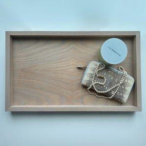 May include: A wooden tray with a light brown finish. The tray contains a white container with the text "VIA MERCATO" and a white and brown patterned wallet with a pearl necklace.