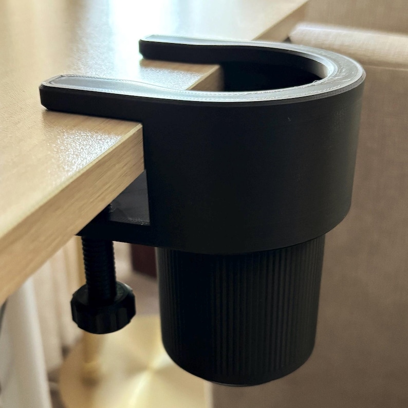 May include: A black clamp-on cup holder with a ribbed design. The holder is attached to a wooden table.
