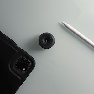 May include: A black phone case with a camera lens cutout and a black round object with a small hole in the center. A white stylus pen is also in the image.