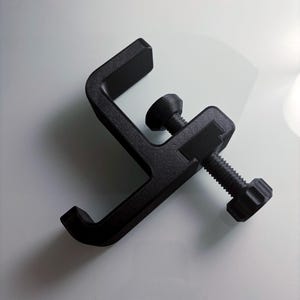 May include: Black plastic clamp with a threaded screw and a knurled knob. The clamp is designed to attach to a surface and hold an object.
