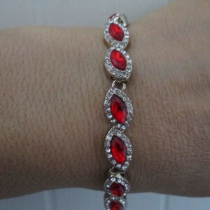 May include: A gold bracelet featuring a line of red marquise-shaped stones set in a clear crystal halo.