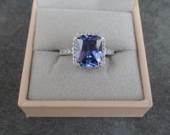 Op de afbeelding: Een zilveren ring met een grote, vierkante, paarse edelsteen omringd door kleinere, heldere edelstenen.