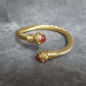 Puede incluir: Un brazalete dorado con un diseño de cable trenzado. Los extremos abiertos del brazalete terminan en tapas metálicas ornamentadas con espirales, cada una con una piedra redonda, lisa y de color rojo anaranjado intenso en el centro.