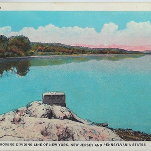 May include: A postcard showing a stone marker on a rocky shore, marking the tri-state border of New York, New Jersey, and Pennsylvania. The marker is inscribed with the words "Tri-State Rock". The postcard features a scenic view of a lake with a forested shoreline and a blue sky with white clouds.