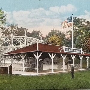 May include: A vintage postcard depicting a wooden roller coaster called "The Blue Streak" at Robison Park in Fort Wayne, Indiana. The coaster is in the background, with a white wooden building in the foreground. The building has a red roof and a sign that reads "The Blue Streak". There is a small American flag flying in the background.