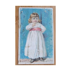 May include: Vintage postcard featuring a young girl in a white dress with a pink sash, finger to her lips. The background is a faded blue with floral accents. Text at the bottom reads "Guess what I got" and "Its awful good."