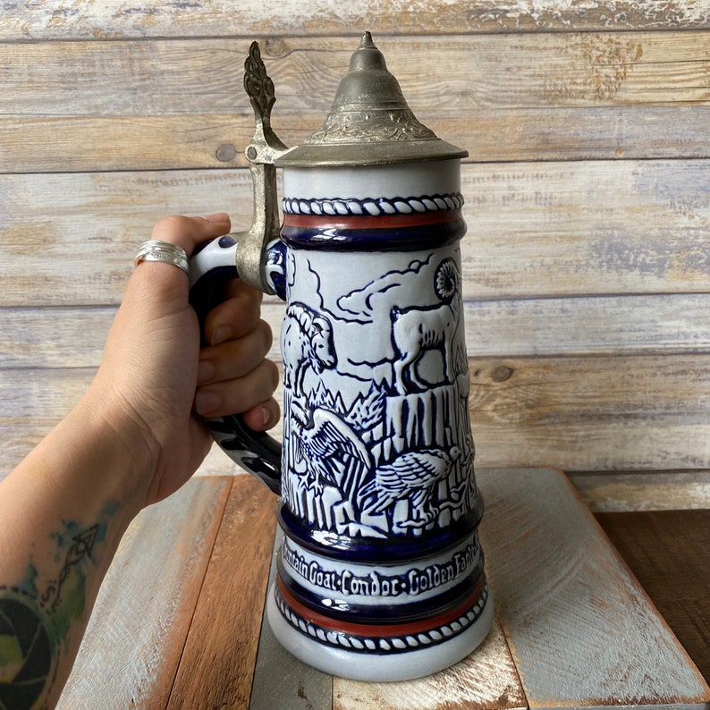 Pu&ograve; includere: Una boccale di birra in ceramica bianca con un design blu e rosso. La boccale presenta una capra di montagna, un condor e un'aquila reale. Il coperchio &egrave; in peltro.