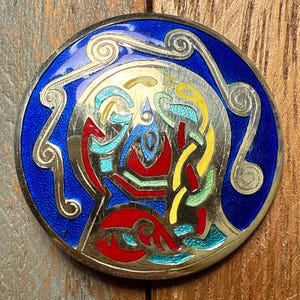 May include: A round, silver-toned brooch with a vibrant enamel design. The brooch features a deep blue border and a central design with red, yellow, green, and blue elements. The design has a Celtic knot style.