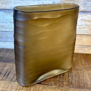 Op de afbeelding: Een rechthoekige, amberkleurige vaas met een textuur, golfachtig patroon. De vaas heeft een afgeronde boven- en onderkant en is gemaakt van een doorschijnend materiaal. De vaas staat op een houten oppervlak.