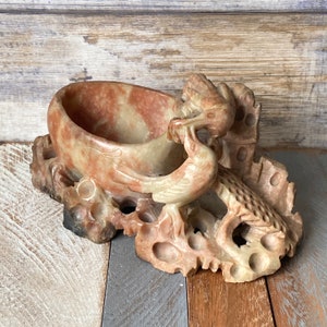 Könnte beinhalten: Eine handgeschnitzte Steinskulptur eines Vogels, der auf einem Ast sitzt, mit einer kleinen Schale, die in den Ast geschnitzt ist. Die Skulptur ist aus einem rötlich-braunen Stein mit weißen und grauen Streifen gefertigt.