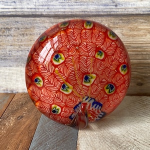 May include: A red glass paperweight featuring a peacock design. The peacock's tail feathers are made of white and blue glass.