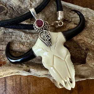 May include: A handcrafted pendant necklace featuring a carved ivory-colored bull skull with black horns. The pendant is attached to a black leather cord and accented with a silver-toned bail and a red gemstone.